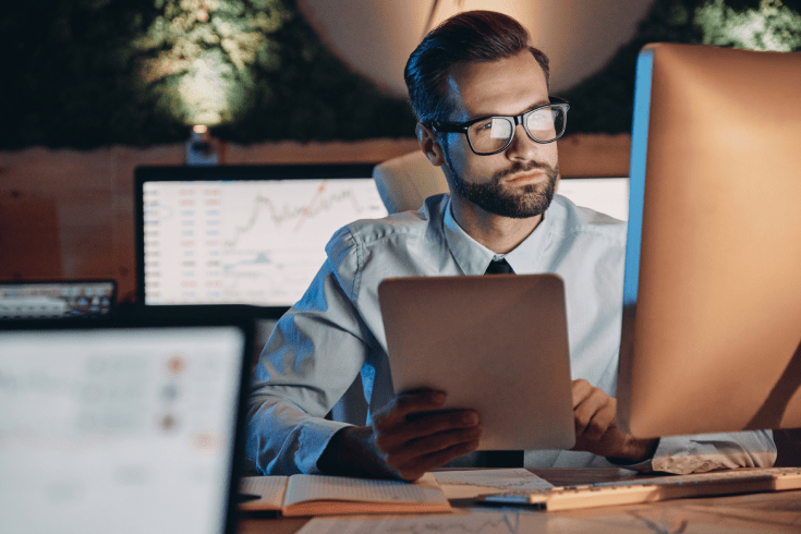 Businessman at desk holding a tablet and looking at a desktop monitor